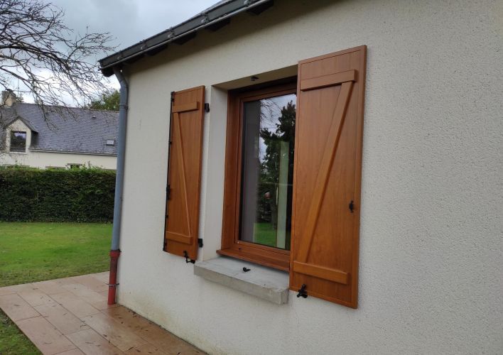 Une fenêtre en bois avec des volets fermés sur un mur clair, entourée d'une pelouse verte.