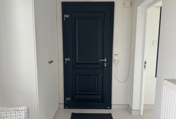 Une porte d'entrée noire au design moderne est entourée d'un cadre mural blanc, avec un sol carrelé clair et un interrupteur électrique à