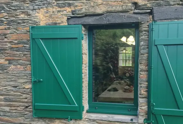 Une fenêtre à volets verts ouverts, encadrée de pierre rustique, offre une vue sur l'intérieur d'une maison chaleureuse.