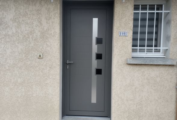 Une porte moderne en acier gris anthracite avec des éléments géométriques en verre, encadrée dans un mur en crépi beige.