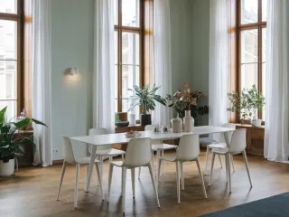 Une salle à manger lumineuse avec une grande table blanche entourée de chaises, baignée de lumière naturelle par de grandes fenêtres et agrémentée