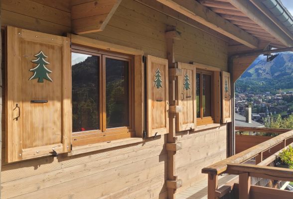 Vue extérieure d'un chalet en bois avec des volets décorés d'arbres, surplombant un paysage montagneux.