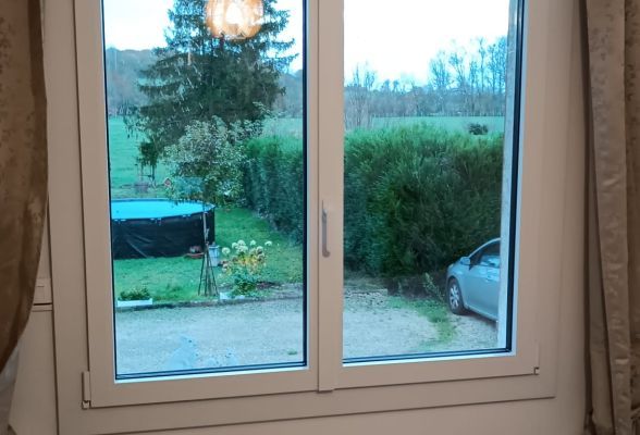 Fenêtre en PVC blanc avec vue sur un jardin verdoyant, un arbre et une piscine extérieure couverte.