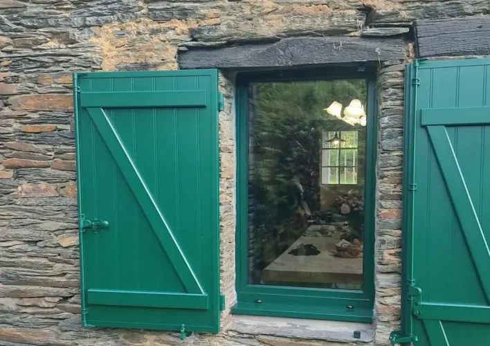 Une fenêtre à volets verts ouverts, encadrée de pierre rustique, offre une vue sur l'intérieur d'une maison chaleureuse.