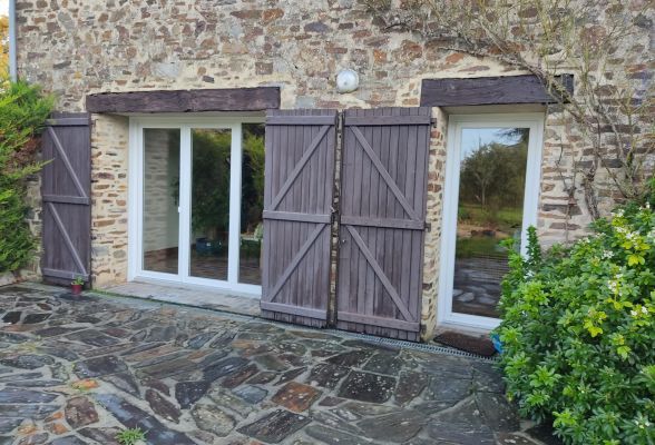 Façade en pierre avec fenêtres à volets en bois, donnant sur une cour en pavés entourée de verdure.