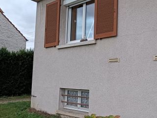 La photo montre un mur extérieur d'une maison avec une fenêtre munie de volets en bois et un petit jardin fleuri en premier plan.