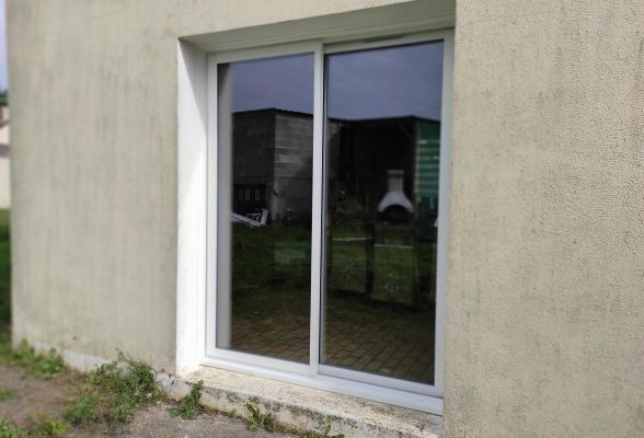 Une fenêtre en verre, intégrée dans un mur beige, reflète un jardin extérieur.