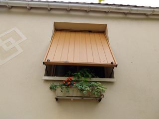 Une fenêtre à volet orange avec une jardinière contenant des plantes vertes et des fleurs rouges est fixée sur une façade beige.