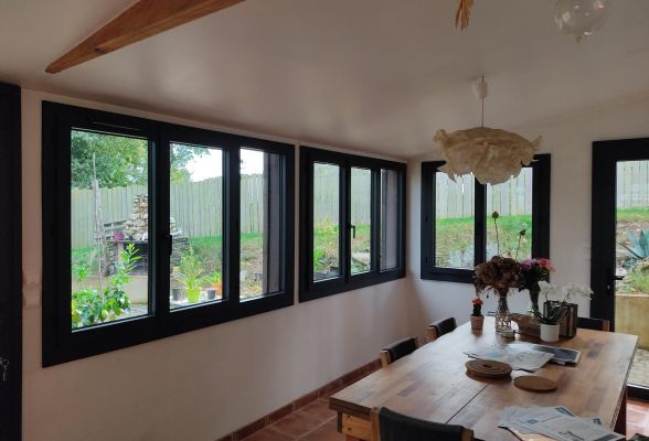 Une salle à manger lumineuse avec une grande table en bois, entourée de fenêtres donnant sur un jardin verdoyant.