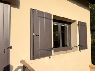 Volet en bois marron fermé sur une maison au crépi beige, baignée de lumière naturelle.