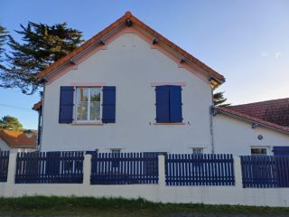 Maison traditionnelle avec volets bleus et toit en tuiles, entourée d'une clôture en bois, sous un ciel dégagé.