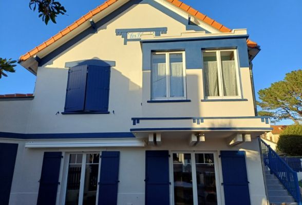 Façade d'une maison traditionnelle avec volets et cadres de fenêtres bleus, illuminée par le soleil.