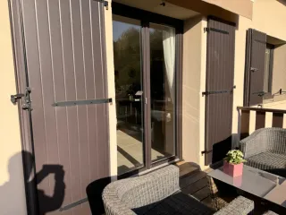 Une terrasse ensoleillée avec des volets en bois foncé, une baie vitrée, et un mobilier d'extérieur en rotin gris.