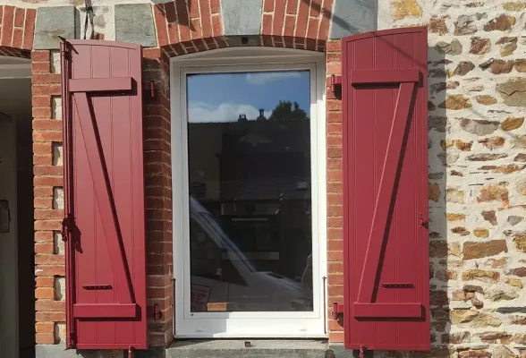 Une fenêtre moderne avec un cadre blanc est entourée de murs en pierre et de volets rouges.