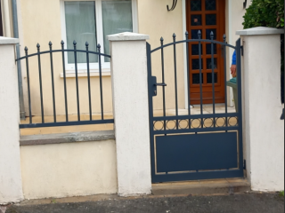 Portail en fer forgé bleu, encadré de piliers blancs, devant une entrée de maison avec une porte en bois vitrée.