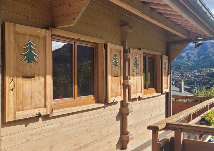 Vue extérieure d'un chalet en bois avec des volets décorés d'arbres, surplombant un paysage montagneux.