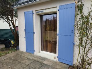 Une porte-fenêtre sur une façade blanchâtre avec des volets bleus et un patio pavé à l'extérieur.