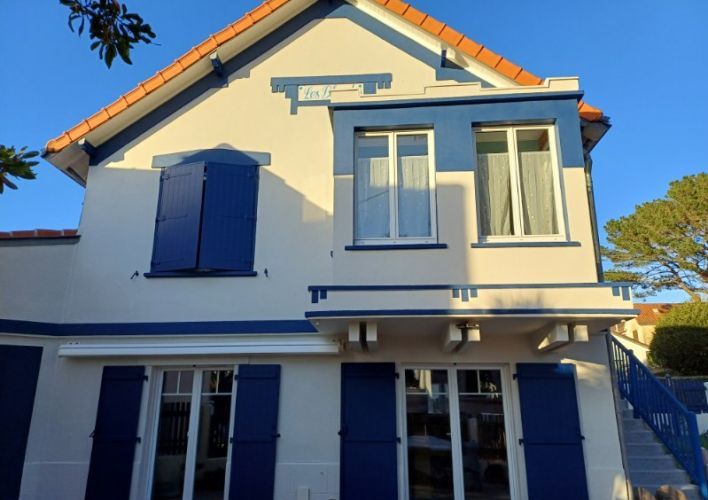 Façade d'une maison traditionnelle avec volets et cadres de fenêtres bleus, illuminée par le soleil.