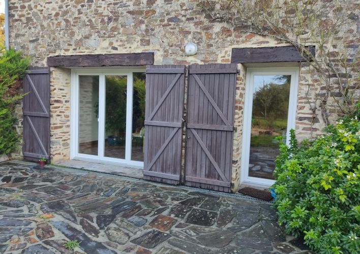 Façade en pierre avec fenêtres à volets en bois, donnant sur une cour en pavés entourée de verdure.