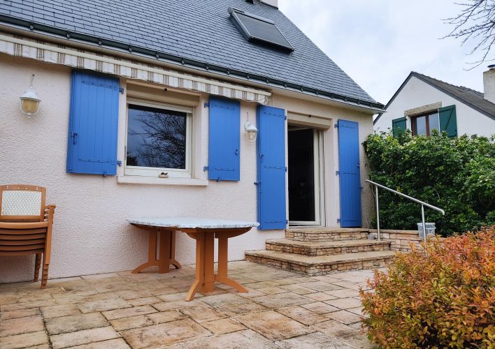 Une charmante terrasse en pierre donnant sur une maison avec des volets bleus et un toit en ardoise, entourée de végétation.