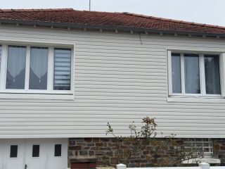 Façade d'une maison avec un revêtement en vinyle blanc, un toit en tuiles rouges et deux grandes fenêtres.