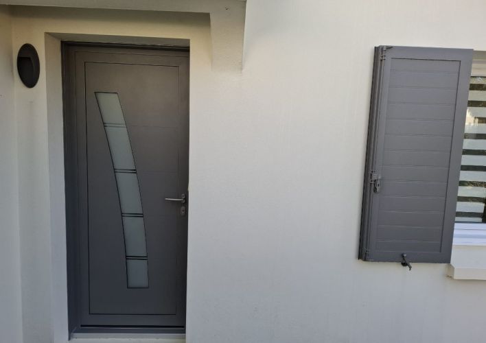 Porte d'entrée moderne en métal gris avec un panneau vitré incurvé, à côté d'un volet assorti sur une façade blanche.