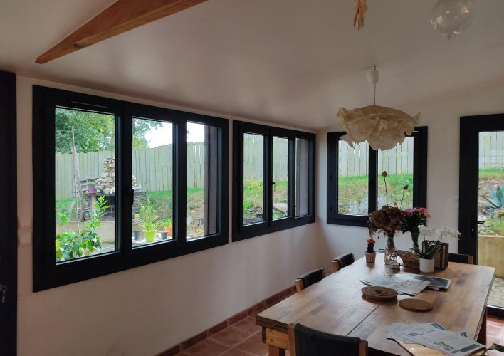 Une salle à manger lumineuse avec une grande table en bois, entourée de fenêtres donnant sur un jardin verdoyant.