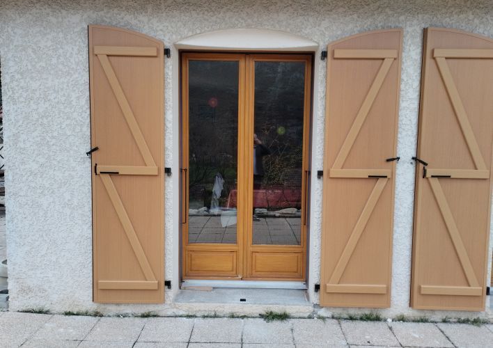 Une porte en bois avec des volets fermés sur une façade en crépi clair.