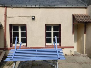 Un banc bleu renversé repose contre un mur beige à côté de deux fenêtres et une porte, dans une petite cour pavée.