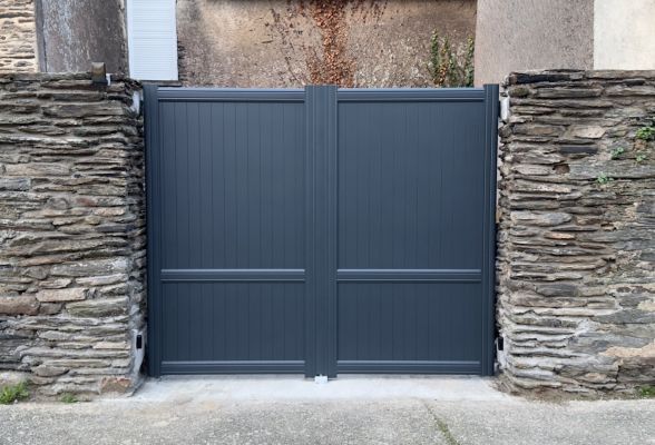 Un portail moderne en métal gris anthracite s'ouvre entre deux piliers en pierre rustique.