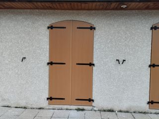 Volet en bois clair avec ferrures noires sur un mur texturé blanc.