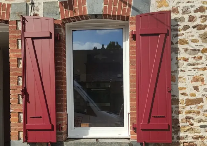 Une fenêtre moderne avec un cadre blanc est entourée de murs en pierre et de volets rouges.