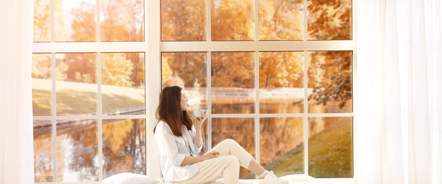 Une femme assise sur un banc devant une grande fenêtre, entourée d'un paysage d'automne, savourant une tasse de café.