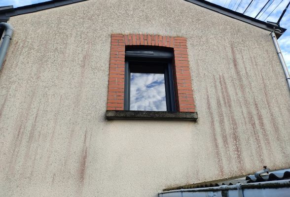 Façade d'une maison avec une fenêtre à encadrement en briques et des traces d'humidité sur le crépi beige.