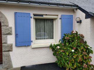 Une fenêtre avec des volets bleus sur une façade claire, accompagnée de verdure et de fleurs devant.