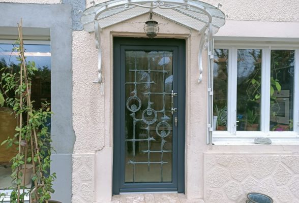 Porte d'entrée élégante avec grille en fer forgé et auvent en métal, encadrée par un mur texturé et des fenêtres.