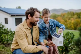 Un homme et une fille sourient en tenant une maquette de maison équipée de panneaux solaires, près d'une maison réelle avec des panneaux solaires